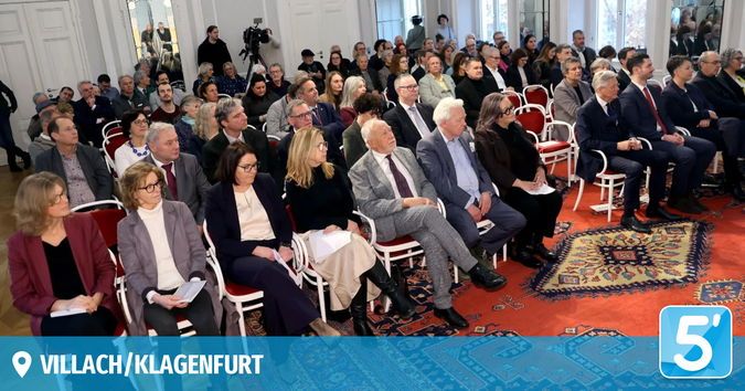 Eine formelle Versammlung mit Menschen in Anzügen und Krawatten, die in Reihen sitzen, einige Papiere halten, auf Stühlen mit einem Teppichboden und einem Schild, das '5' in der Ecke steht.