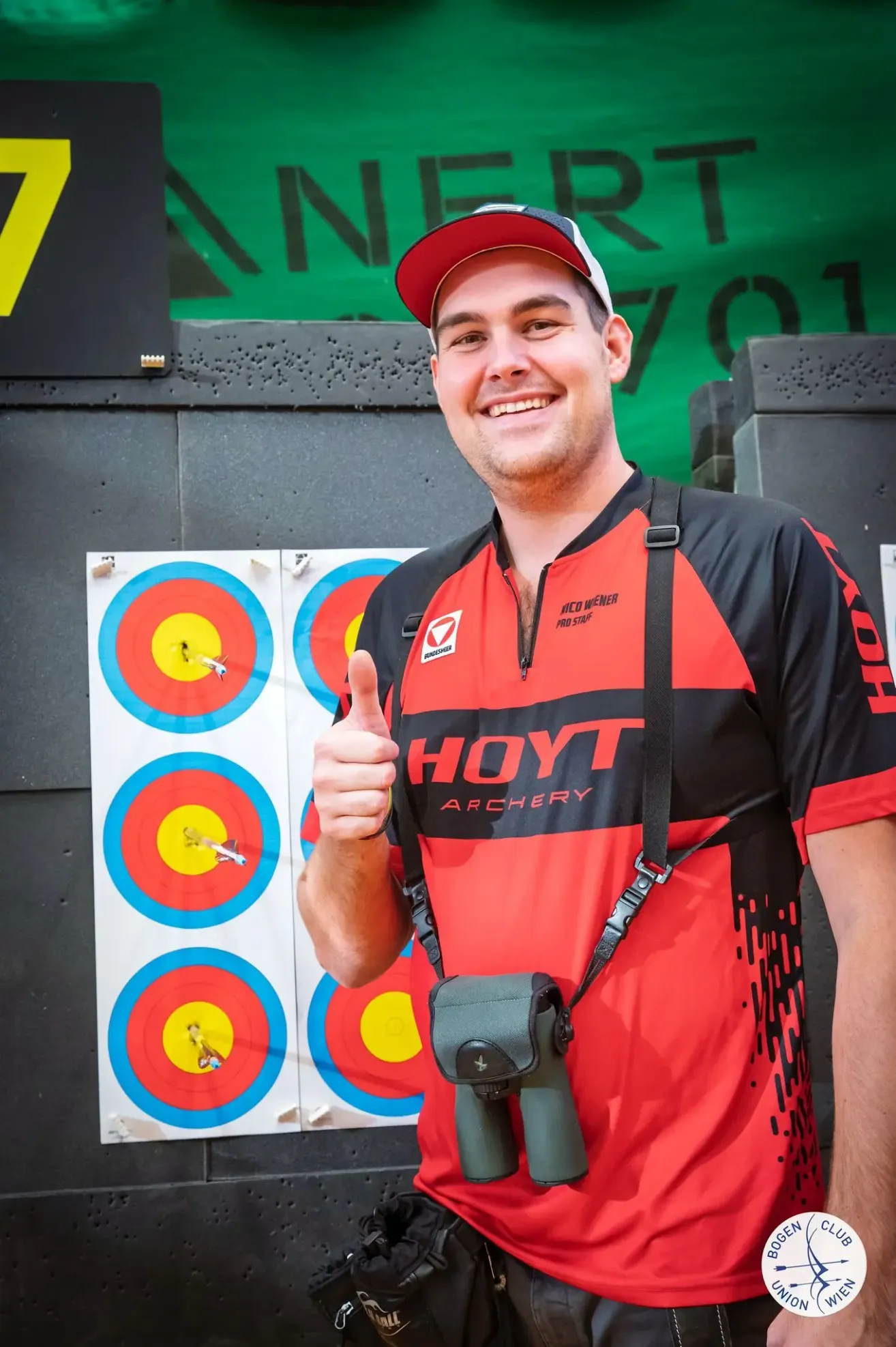 A smiling man in a red and black shirt, wearing a cap, gives a thumbs up in front of a wall with archery targets.