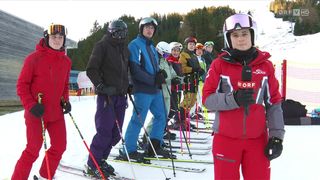 A group of skiers are standing in a line, wearing winter gear and holding ski poles. They are in a snowy area with trees in the background.