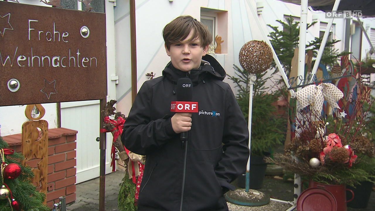 Ein junger Junge in einem schwarzen Jacke mit ORF-Logo steht vor einem Haus und hält ein Mikrofon. Hinter ihm sind Pflanzen und ein Holzschild mit dem Wort 'ten'.