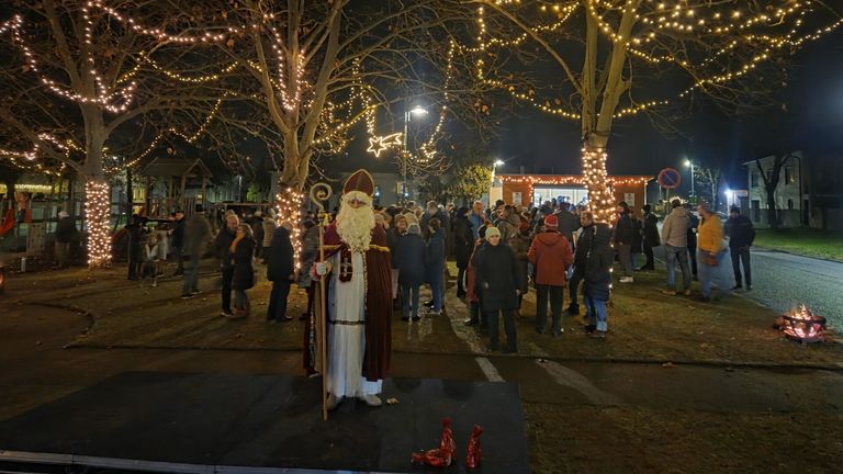 Bei Nacht steht ein als Weihnachtsmann verkleideter Mann auf einer Plattform, umgeben von Menschen in einem festlichen Außenbereich mit Bäumen, die mit Lichtern geschmückt sind.