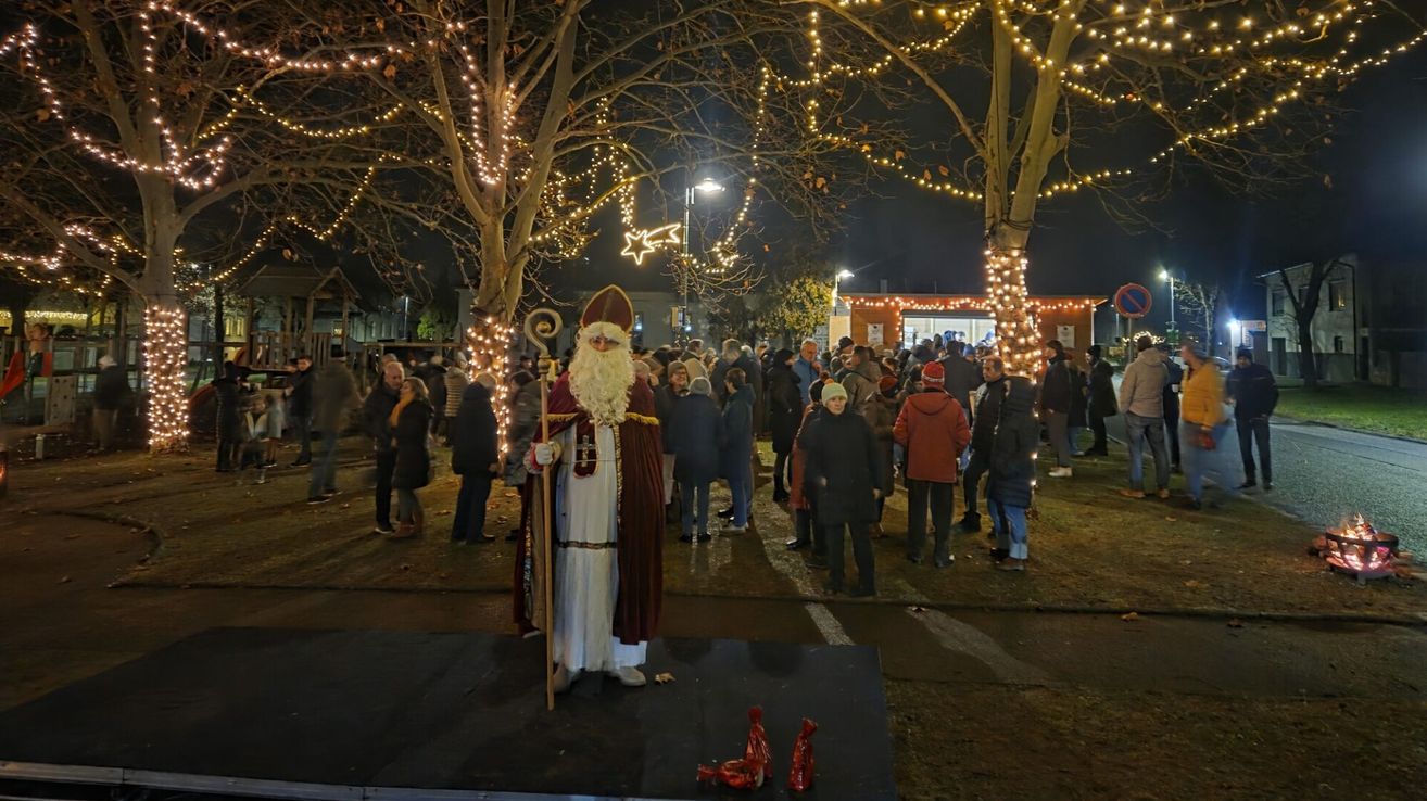 Bei Nacht steht ein als Weihnachtsmann verkleideter Mann auf einer Plattform, umgeben von Menschen in einem festlichen Außenbereich mit Bäumen, die mit Lichtern geschmückt sind.