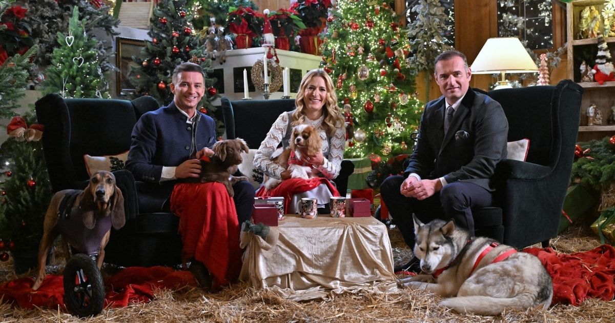 Drei Menschen sitzen mit Hunden auf einer Couch, umgeben von Weihnachtsdekoration und Geschenken. Der Mann links hält einen Hund, während die Frau in der Mitte einen anderen hält. Ein Husky liegt auf dem Boden.