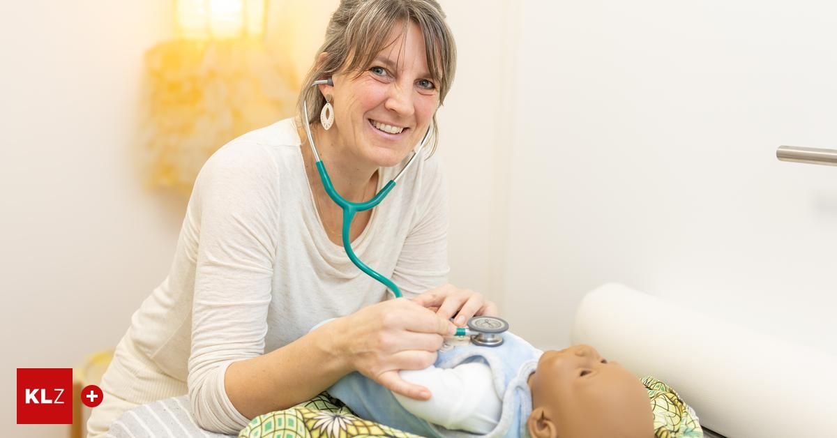 Eine lächelnde Frau mit einem Stethoskop um den Hals hält eine Baby-Puppe. Die Puppe liegt auf einem Bett mit einer bunten Decke.