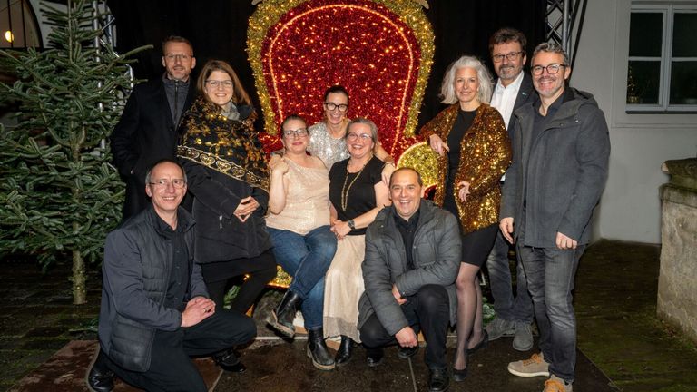 Eine Gruppe von Menschen posiert für ein Foto vor einem dekorierten Stuhl, wahrscheinlich für eine festliche Veranstaltung. Der Stuhl ist mit rotem und goldenem Glitzer verziert.