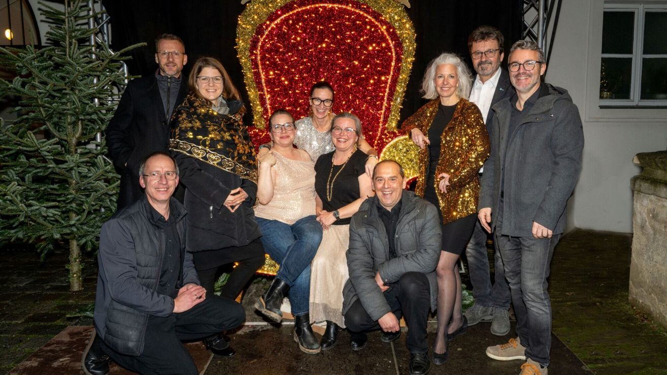 Eine Gruppe von Menschen posiert für ein Foto vor einem dekorierten Stuhl, wahrscheinlich für eine festliche Veranstaltung. Der Stuhl ist mit rotem und goldenem Glitzer verziert.