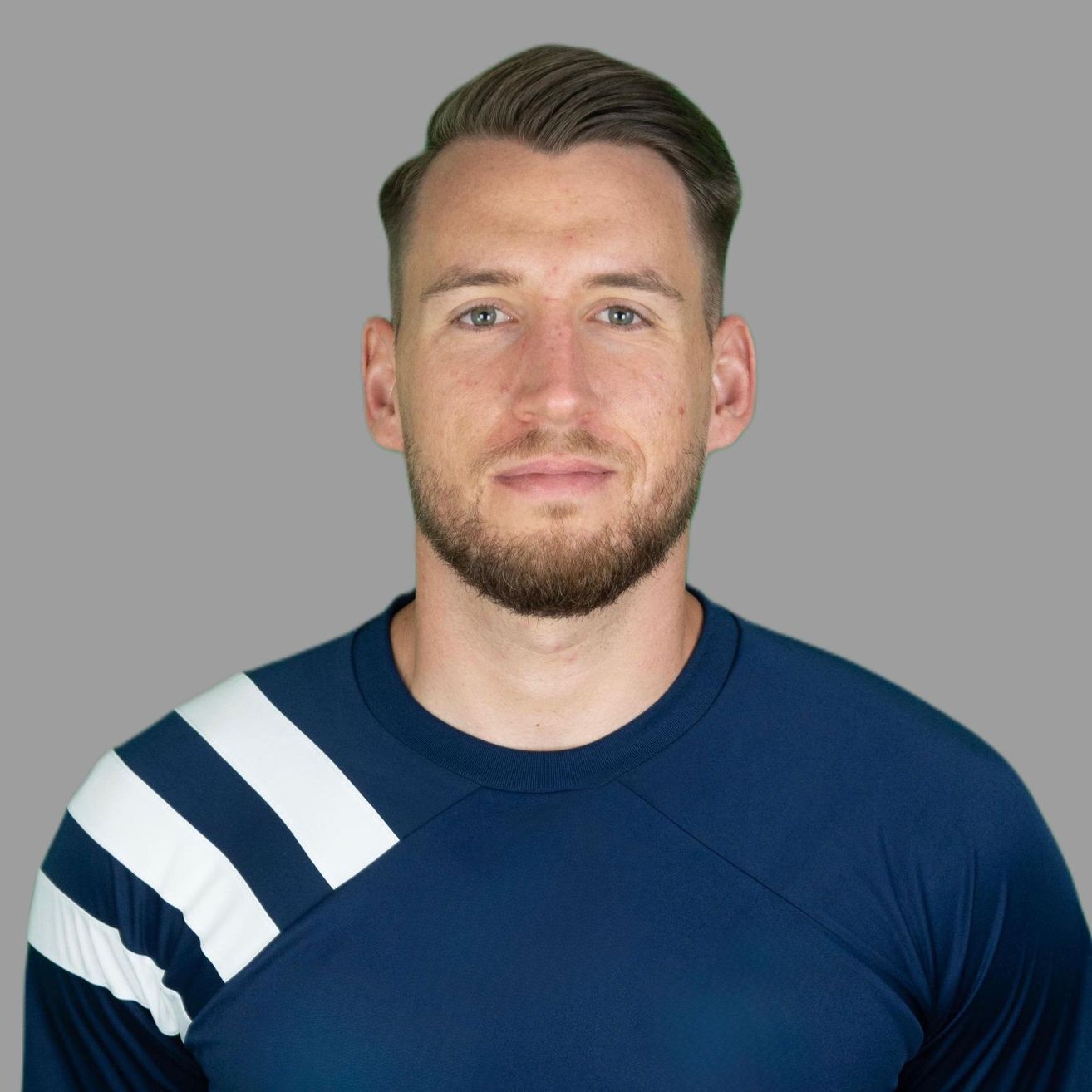 Ein Mann mit Bart trägt ein dunkelblaues T-Shirt mit weißen Streifen an den Ärmeln. Er schaut direkt in die Kamera.