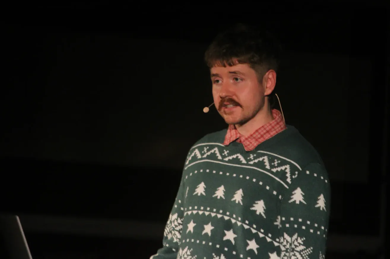 Ein Mann mit einem Mikrofon und einem grünen Pullover mit Weihnachtsbaum-Muster schaut nach vorne.