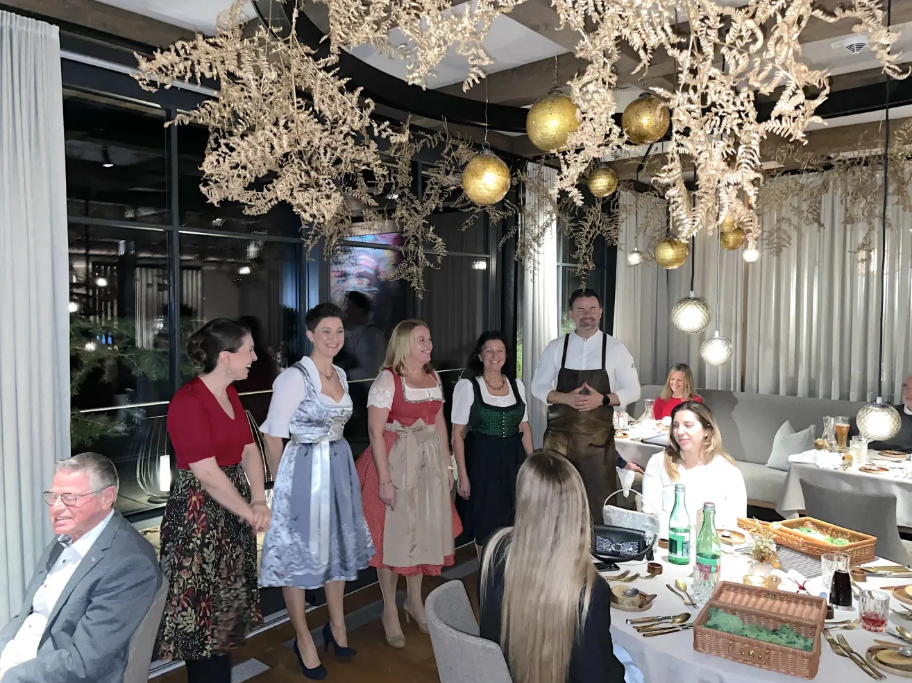 Mehrere Frauen in traditioneller Kleidung stehen und lächeln, während ein Mann in einer Schürze vor ihnen steht. Der Raum ist mit goldenen Ornamenten dekoriert.