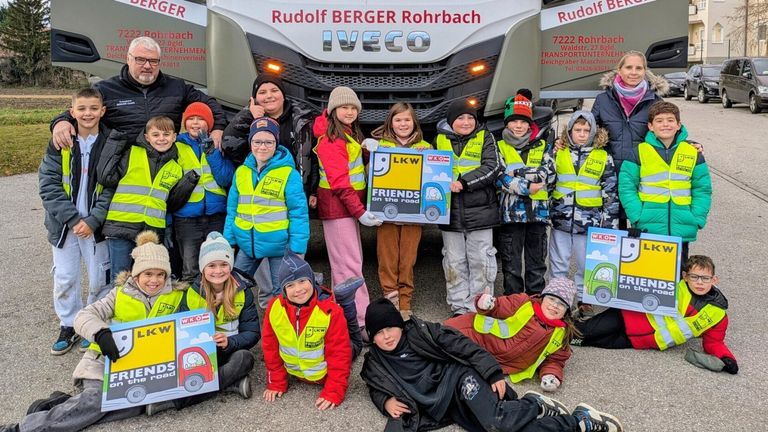 Kinder und Erwachsene in Warnwesten posieren mit einem Schild, das 'Freunde auf der Straße' vor einem IVECO-LKW steht.