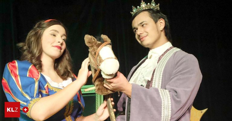 Ein Mann und eine Frau in Kostümen, wahrscheinlich für ein Theaterstück, halten ein Stoffpferd. Der Mann trägt eine Krone und lächelt, während die Frau ihn ansieht.