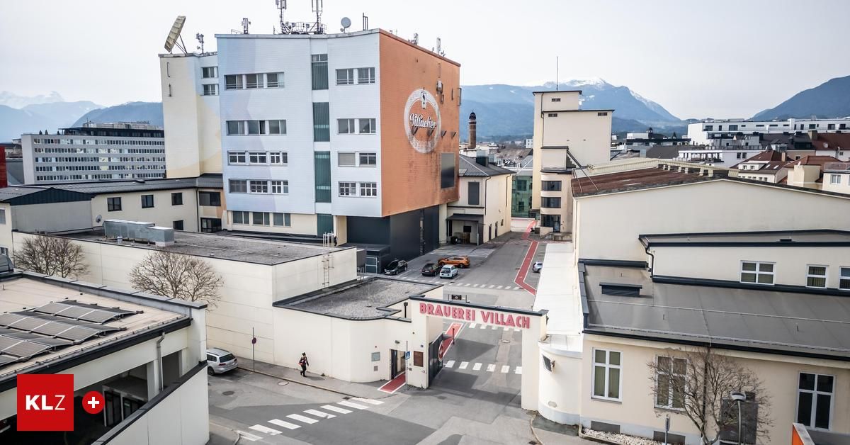 Ein Luftbild von Brauerei Villach zeigt das Hauptgebäude mit einem Logo, mehrere geparkte Autos, einen Fußgänger und Berge in der Ferne.