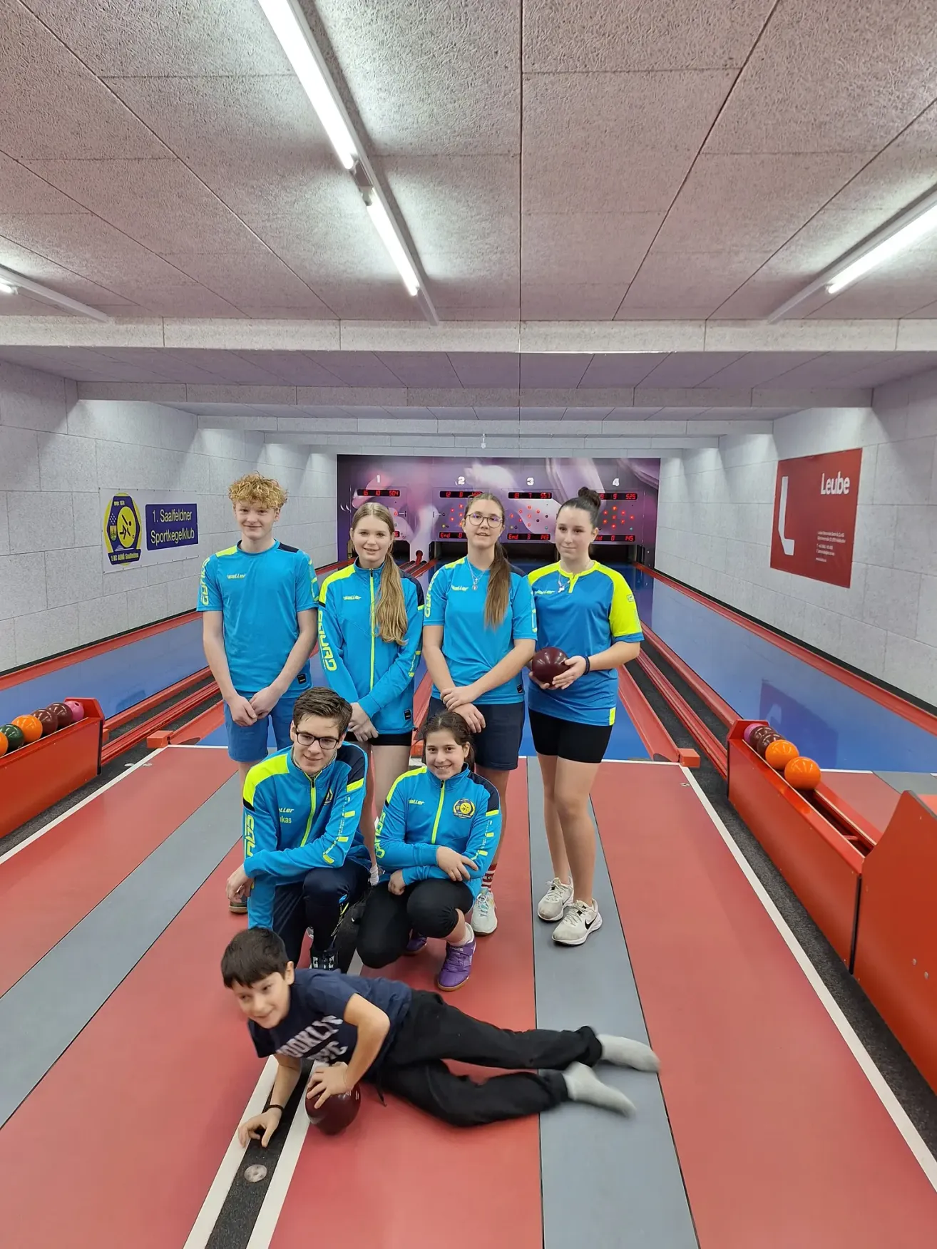 Eine Gruppe junger Leute in passenden blauen Uniformen posiert für ein Foto in einer Bowlingbahn. Die jungen Leute stehen und knien in verschiedenen Posen.