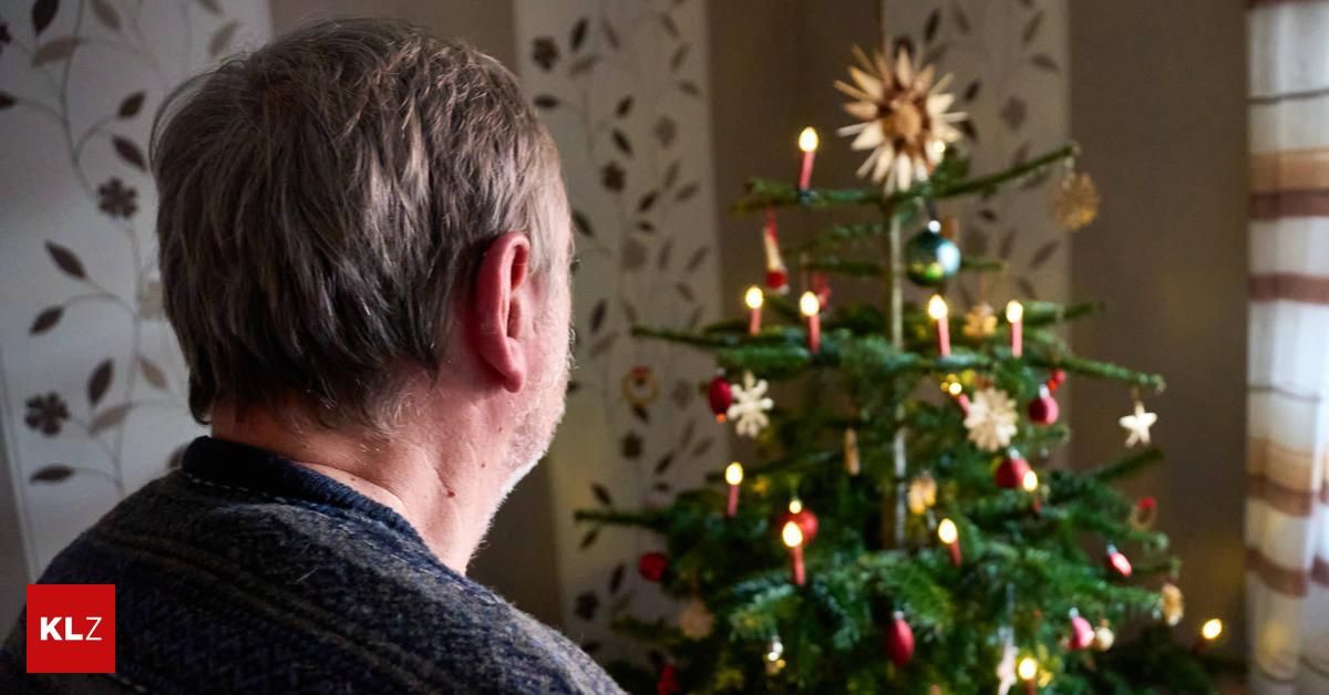 Ein älterer Mann betrachtet einen geschmückten Weihnachtsbaum mit brennenden Kerzen, Ornamenten und einer Sonnenblume obenauf.