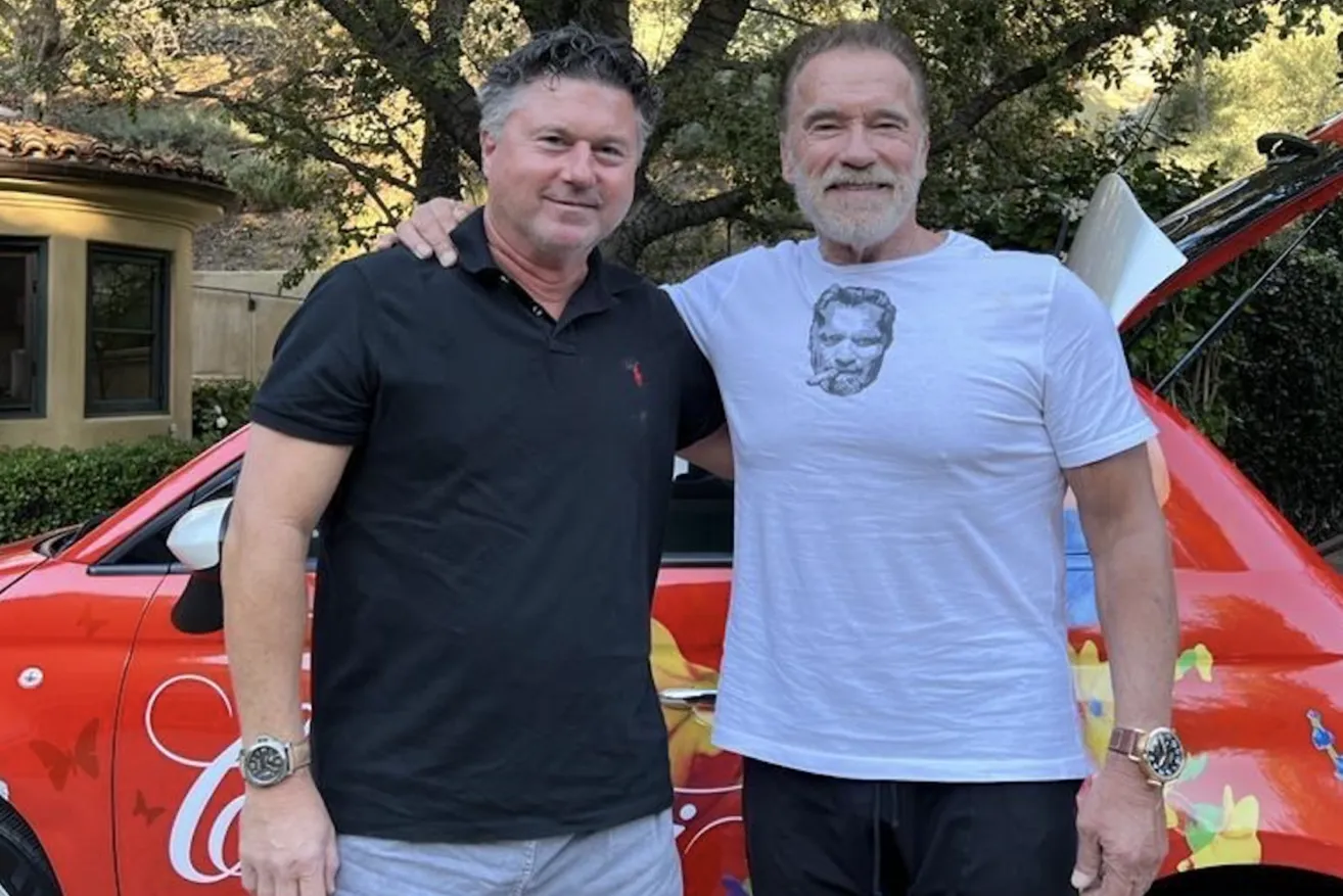 Zwei Männer stehen nebeneinander vor einem roten Auto, einer trägt ein schwarzes Poloshirt und der andere ein weißes T-Shirt mit dem Bild eines bärtigen Mannes.