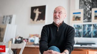 Ein älterer Mann mit Brille und schwarzem Pullover sitzt in einem Kunststudio, umgeben von Kunstwerken an den Wänden.