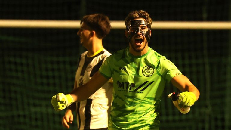 Ein Fußballspieler in einem grünen Trikot feiert in der Nähe des Tores. Ein maskierter Torwart mit Handschuhen steht vor ihm. Ein weiterer Spieler in einem weißen Trikot beobachtet. Die Umgebung ist nachts.