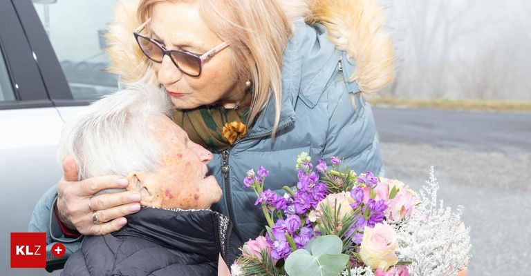 Eine ältere Frau mit Brille und Schal wird von einer anderen Frau in einer blauen Jacke mit Pelzkragen getröstet. Sie halten einen Blumenstrauß.