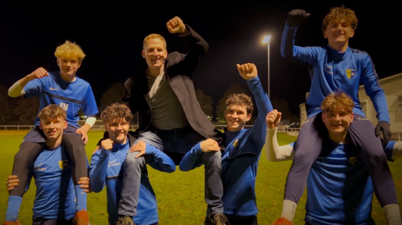 Eine Gruppe junger Männer feiert auf einem Feld bei Nacht. Ein Mann sitzt auf den Schultern von zwei anderen, alle tragen blaue Trikots.