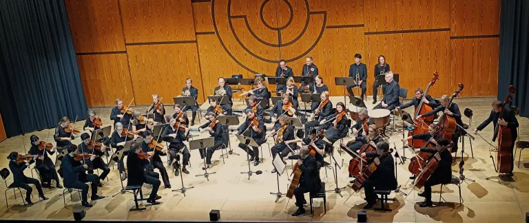 Ein Symphonieorchester auf der Bühne, Musiker spielen verschiedene Instrumente wie Violinen, Celli und Blechbläser, mit einem hölzernen Hintergrund und kreisförmigem Design.