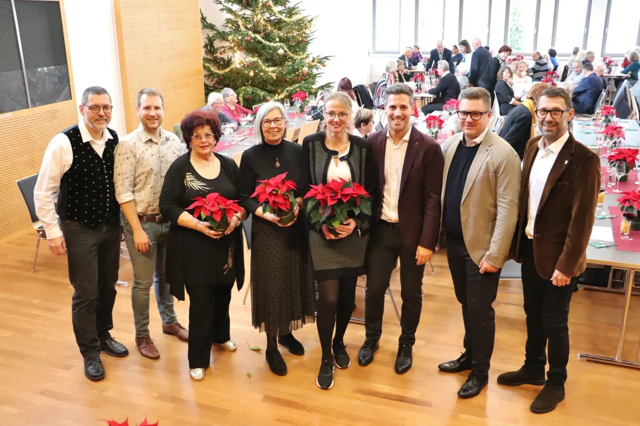 Eine Gruppe von Menschen steht lächelnd zusammen und hält Blumen in der Hand, vor einem Weihnachtsbaum. Die Umgebung ist festlich, mit weiteren Personen, die hinter ihnen sitzen und stehen.