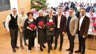 Eine Gruppe von Menschen steht lächelnd zusammen und hält Blumen in der Hand, vor einem Weihnachtsbaum. Die Umgebung ist festlich, mit weiteren Personen, die hinter ihnen sitzen und stehen.