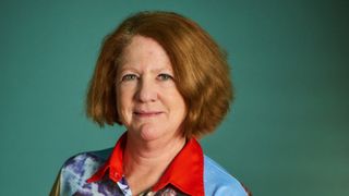 A woman with short brown hair and green eyes is smiling slightly. She wears a blue and red collared shirt against a green background.