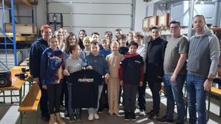 Eine Gruppe von Kindern und Erwachsenen posiert für ein Foto in einem Lagerhaus, einige halten ein schwarzes T-Shirt mit dem Wort 'FEUERWEHR' darauf.