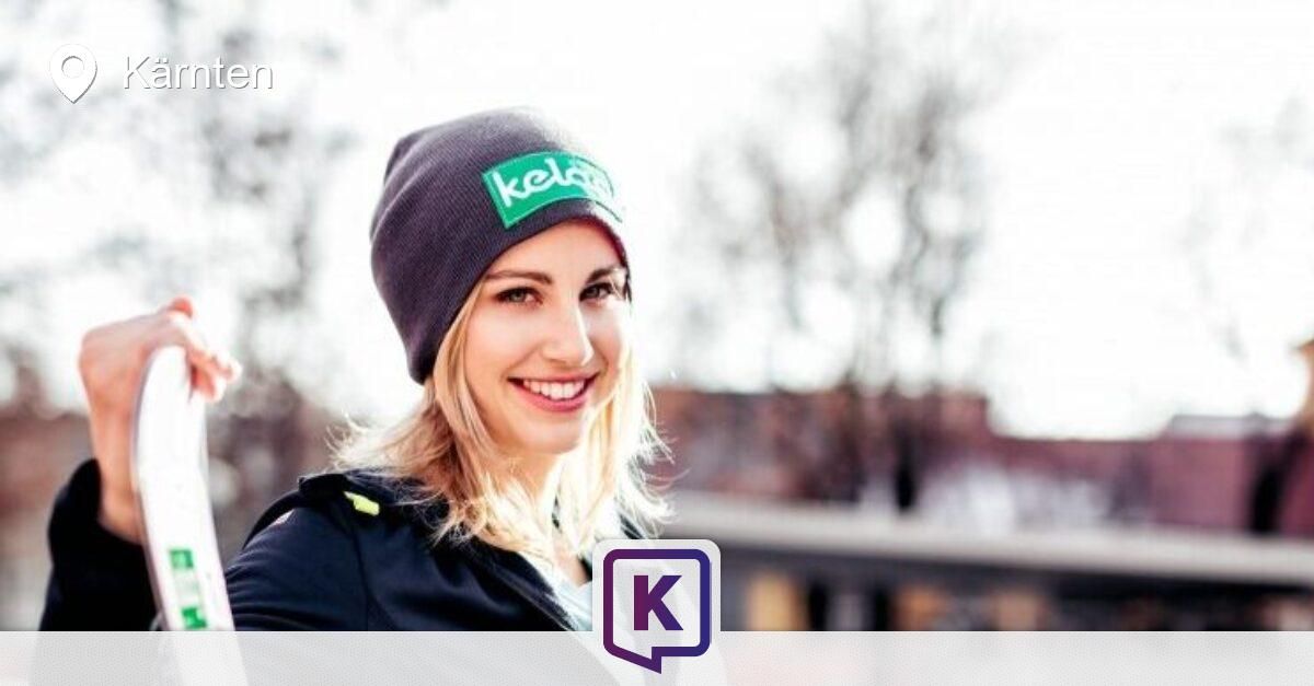 Eine Frau in einer schwarzen Jacke und einer Mütze mit dem Wort 'kela' darauf lächelt und steht vor einem verschwommenen Hintergrund.