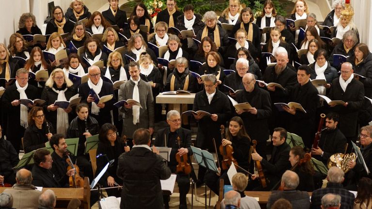 Ein großer Chor, gekleidet in schwarze und weiße Schals, liest aus Musikbüchern und spielt Violinen, mit einem Dirigenten vorn.