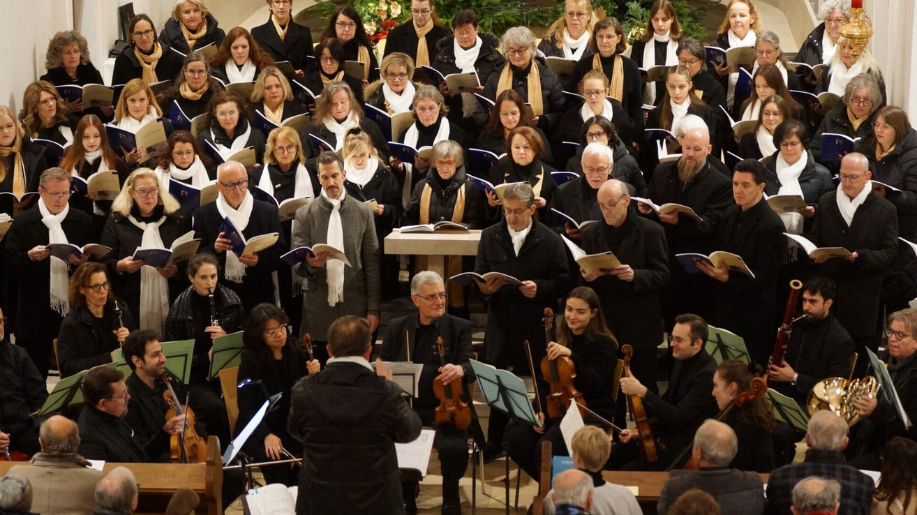 Ein großer Chor, gekleidet in schwarze und weiße Schals, liest aus Musikbüchern und spielt Violinen, mit einem Dirigenten vorn.
