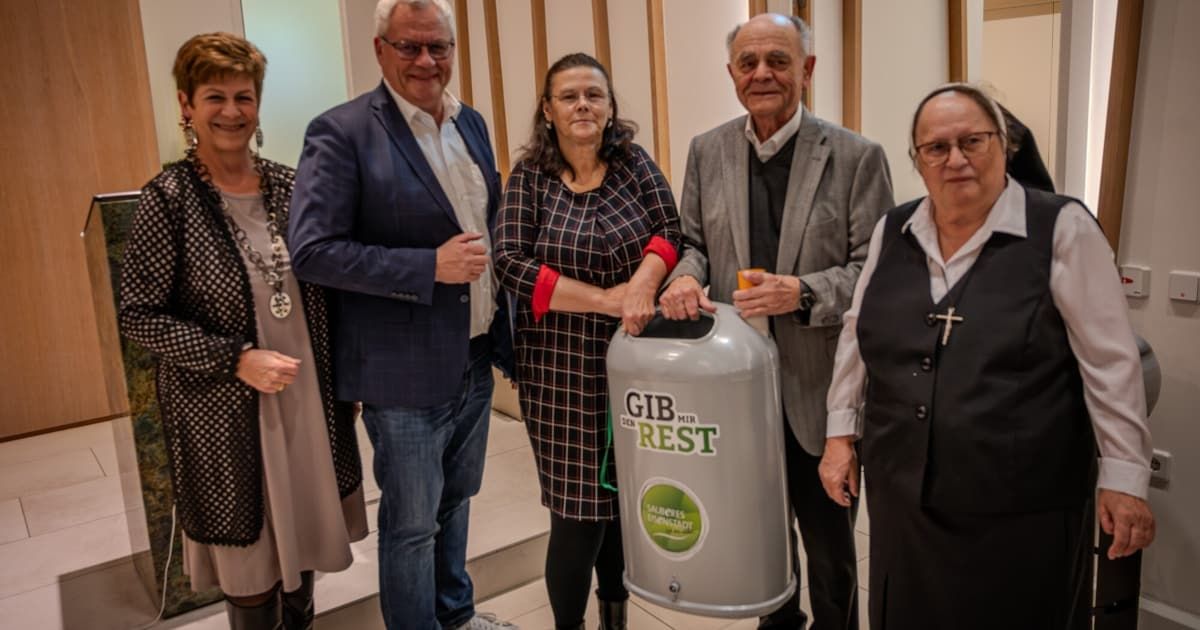 Fünf Menschen stehen in einem Raum zusammen. Die zentrale Figur ist eine Frau, die einen Recyclingbehälter hält. Der Behälter hat ein Etikett, das 'GIB MIR REST' besagt. Die Gruppe scheint für ein Foto zu posieren.