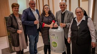 Fünf Menschen stehen in einem Raum zusammen. Die zentrale Figur ist eine Frau, die einen Recyclingbehälter hält. Der Behälter hat ein Etikett, das 'GIB MIR REST' besagt. Die Gruppe scheint für ein Foto zu posieren.