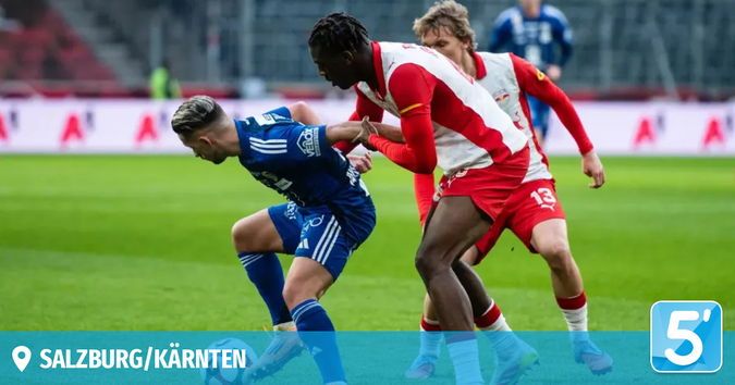 Drei Fußballspieler auf einem grünen Feld, zwei in Blau und einer in Rot, spielen um den Ball. Ein Logo für Salzburg/Kärnten ist unten links.
