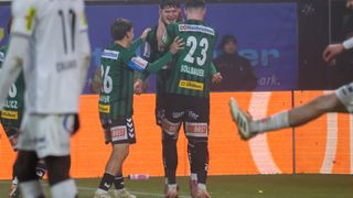 Drei Fußballspieler in grünen Trikots feiern auf dem Feld. Einer trägt das Trikot Nummer 23, ein anderer 26. Im Hintergrund ein orangefarbener Hintergrund mit Sponsorenlogos.