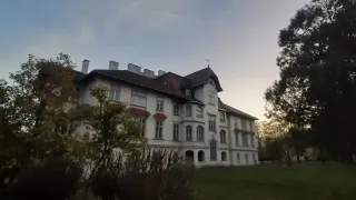 Ein großes, weißes Gebäude mit mehreren Fenstern und einem braunen Dach, umgeben von einem Rasen und Bäumen, unter einem blauen Himmel.