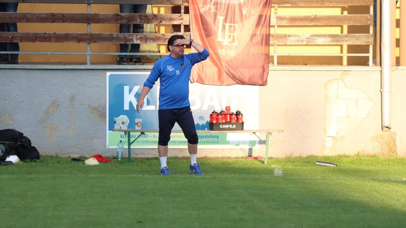 Ein Trainer in einem blauen Trikot steht auf einem Fußballfeld und schaut die Spieler an. Hinter ihm steht eine Holzbank mit Flaschen, ein Banner und eine Wand mit einem blauen Schild.