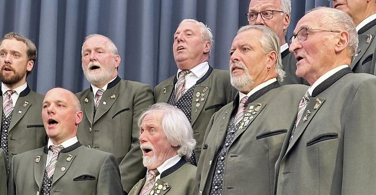 Eine Gruppe älterer Männer in passenden grünen Anzügen und Krawatten, möglicherweise ein Chor, singen auf der Bühne.