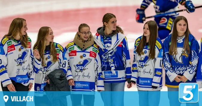 Sieben Frauen in blau-weißen Eishockeytrikots stehen in einer Reihe, möglicherweise auf einer Eisbahn. Sie scheinen für ein Foto zu posieren. Im Hintergrund ist eine verschwommene Figur mit einem Hockeyschläger zu sehen.