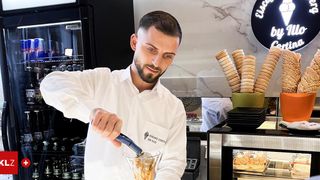 Ein Mann in einem weißen Hemd bereitet in einer modernen Küche mit Marmorarbeitsplatten ein Getränk in einem Glas mit einem Löffel zu.