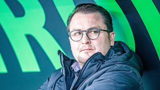 Ein Mann in einer blauen Jacke mit Kapuze und einer Brille steht vor einem grün-blauen Hintergrund.