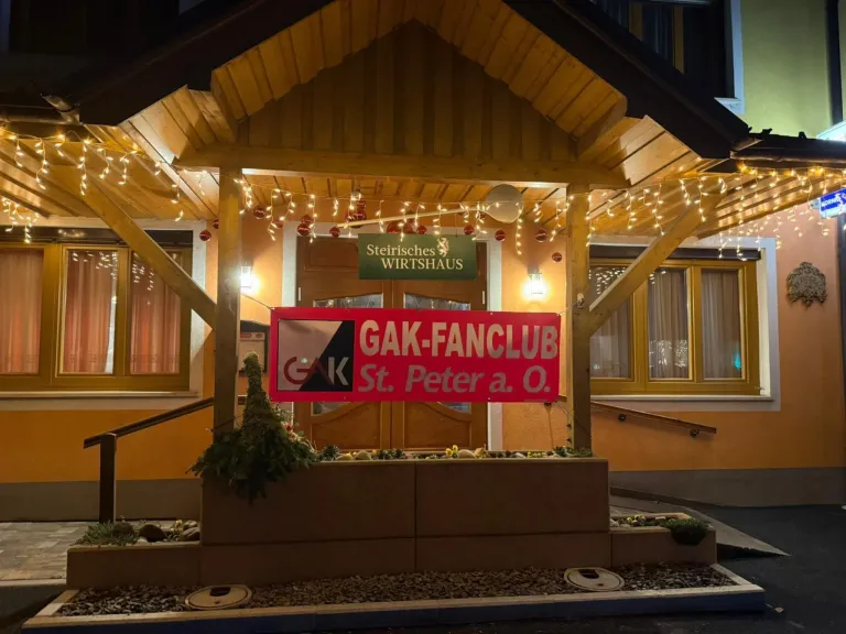 Ein Restaurant mit dem Schild 'GAK-FANCLUB St. Peter a. O.' ist beleuchtet und hat eine Pflanze davor.
