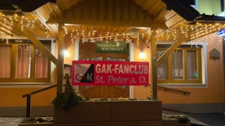Ein Restaurant mit dem Schild 'GAK-FANCLUB St. Peter a. O.' ist beleuchtet und hat eine Pflanze davor.