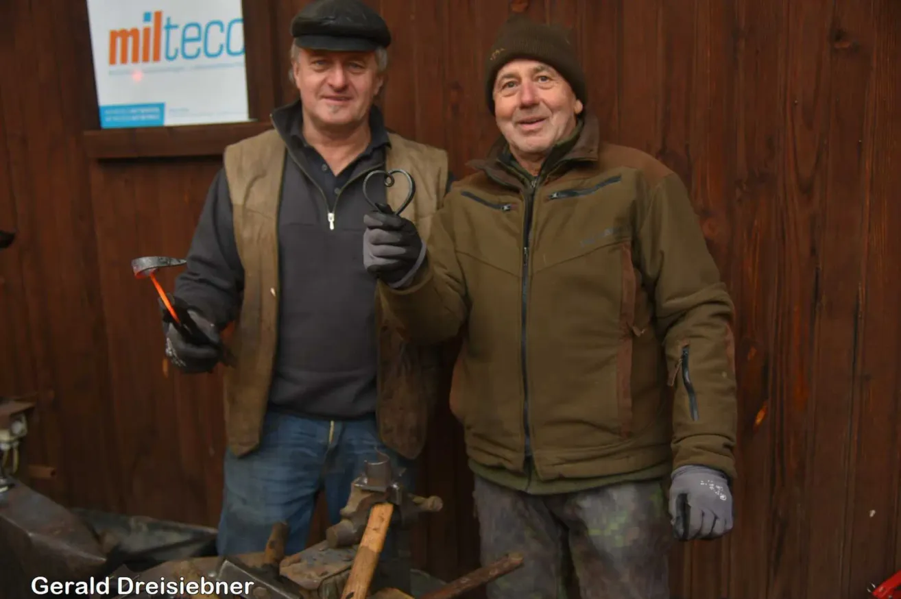 Zwei Männer stehen lächelnd vor einer Holzwand. Einer hält eine Metallherzform. Hinter ihnen sind Werkzeuge und ein Schild mit der Aufschrift 'lteco' zu sehen.