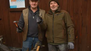 Zwei Männer stehen lächelnd vor einer Holzwand. Einer hält eine Metallherzform. Hinter ihnen sind Werkzeuge und ein Schild mit der Aufschrift 'lteco' zu sehen.