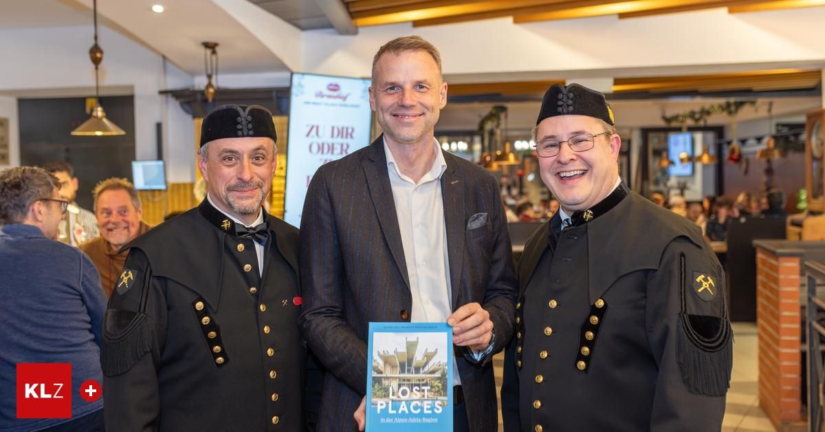Drei Männer stehen zusammen in Innenräumen. Der Mann in der Mitte hält ein Buch mit dem Titel 'Lost Places'. Die Männer sind in übereinstimmenden Uniformen mit Medaillen gekleidet. Der Hintergrund zeigt ein Werbeboard.