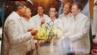 Fünf Männer in weißen Mänteln versammeln sich um einen Tisch mit einem Blumenstrauß und Kerzen. Sie scheinen ein Ereignis zu feiern, vielleicht ein Feiertag. Eine Unterschrift ist auf dem Strauß signiert.