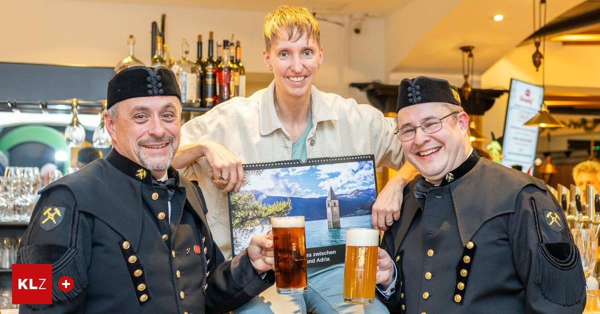 Drei Personen, zwei in Uniform und eine im weißen Hemd, posieren mit Biergläsern in einer Bar. Dahinter zeigt ein Kalender eine Burg und Berge.