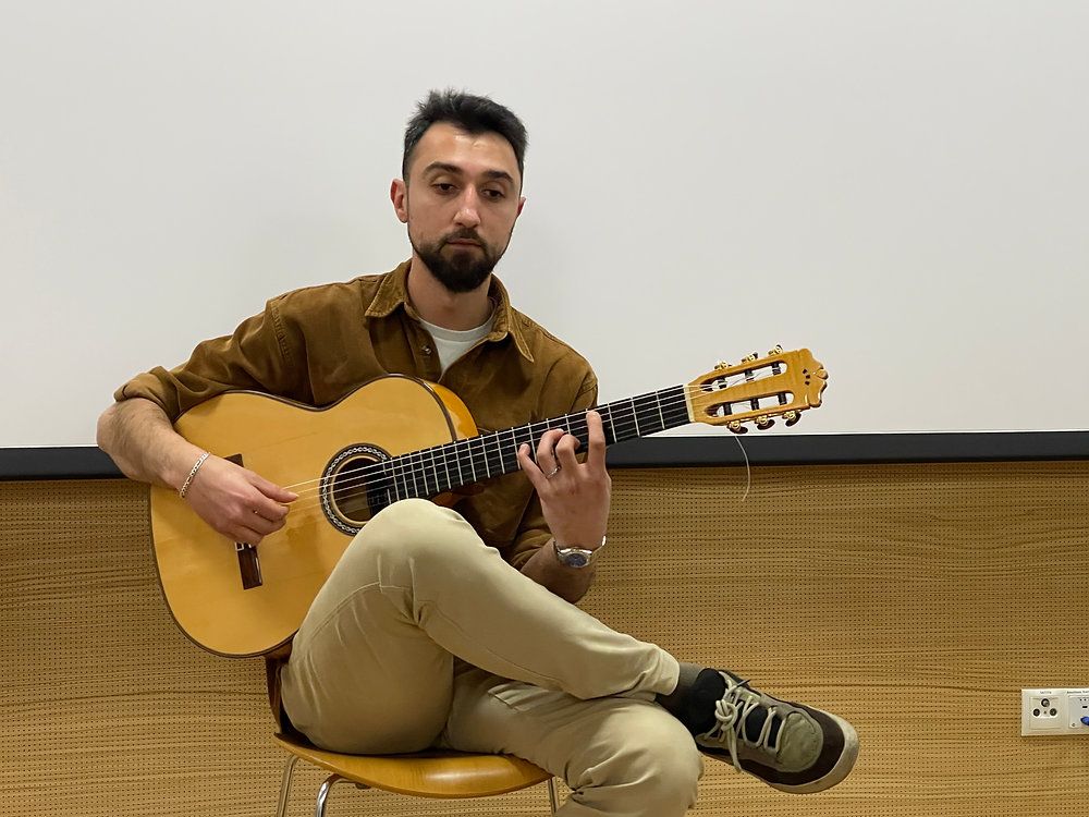 Ein Mann sitzt auf einem Stuhl und spielt eine klassische Gitarre. Er trägt ein braunes Hemd, Khakihosen und Turnschuhe. Er sieht konzentriert aus, mit seinen Augen auf den Gitarrensaiten.