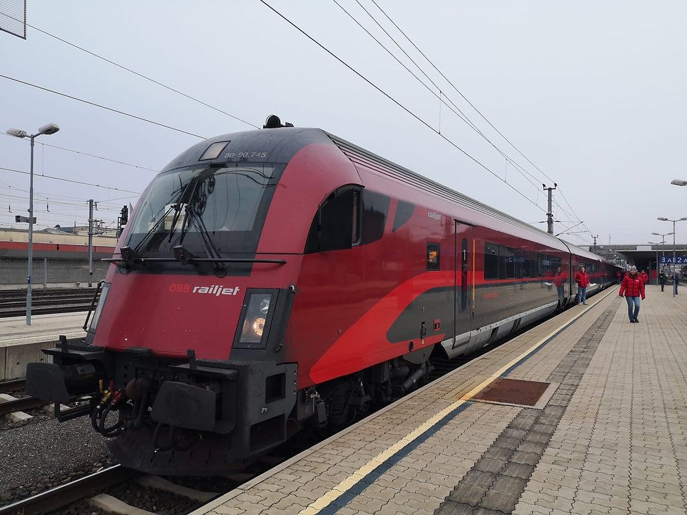 Ein roter Railjet-Zug mit der Nummer 80-96-745 steht auf dem Bahnsteig eines Bahnhofs. Eine Person in einer roten Jacke geht auf dem Bahnsteig.